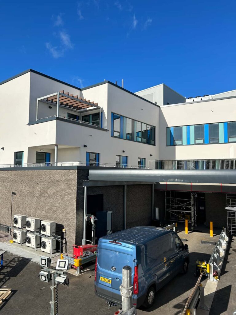 Large white modern looking building with lots of windows in an l-shape and a parking bay with a blue van in it